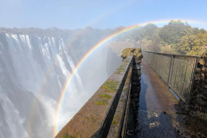 Weg entlang der Victoriafälle mit einem leuchtenden Regenbogen im Nebel.